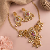 Gold-toned traditional jewelry set featuring a statement necklace and matching earrings with pearl-like beads, pink stone accents, and clear rhinestone details, arranged on a soft pink background with burlap and a flower prop.