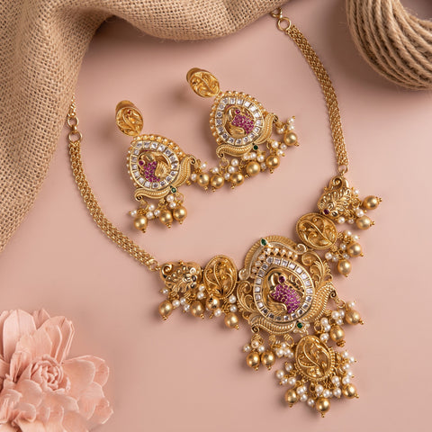Gold-toned traditional jewelry set featuring a statement necklace and matching earrings with pearl-like beads, pink stone accents, and clear rhinestone details, arranged on a soft pink background with burlap and a flower prop.