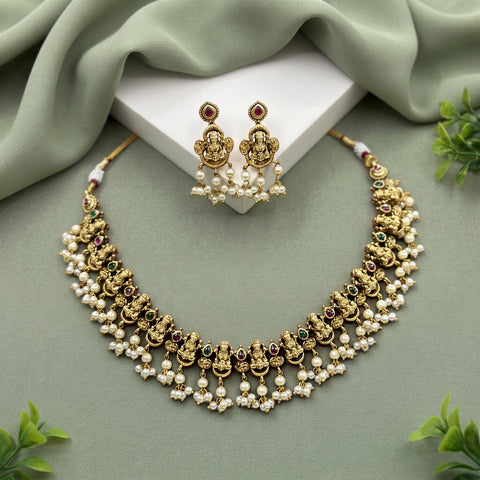Gold-toned traditional Indian jewelry set featuring a necklace and matching earrings with Ganesha motifs, white pearl-like beads, and small green and red stone accents, arranged on a sage green draped fabric background with a white display block.