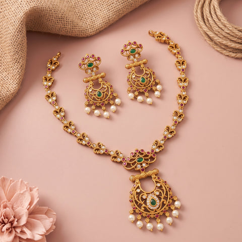 Gold-toned traditional Indian jewelry set featuring a necklace, earrings, and a pendant, embellished with green stones, red stones, and white pearls, arranged on a dusty pink background with decorative burlap and a pink flower.