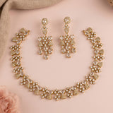 Gold-toned bridal jewelry set featuring a matching necklace, bracelet, and drop earrings, embellished with clear rhinestones on a soft pink background with a beige fabric corner and a pink rose.