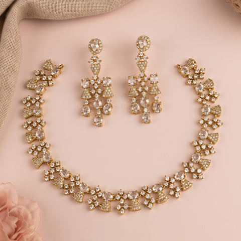 Gold-toned bridal jewelry set featuring a matching necklace, bracelet, and drop earrings, embellished with clear rhinestones on a soft pink background with a beige fabric corner and a pink rose.