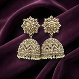 A pair of ornate gold-toned Indian jhumka earrings decorated with clear stones and small pearl-like beads, displayed on draped deep purple fabric.