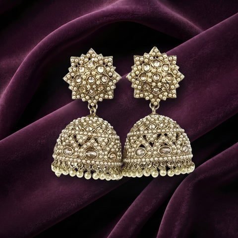 A pair of ornate gold-toned Indian jhumka earrings decorated with clear stones and small pearl-like beads, displayed on draped deep purple fabric.