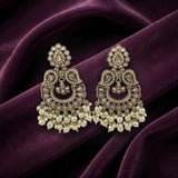 A pair of ornate Indian jhumka earrings with intricate gold-toned filigree, clear stones, and dangling pearl clusters, displayed on draped deep purple velvet fabric.