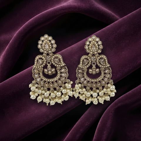 A pair of ornate Indian jhumka earrings with intricate gold-toned filigree, clear stones, and dangling pearl clusters, displayed on draped deep purple velvet fabric.