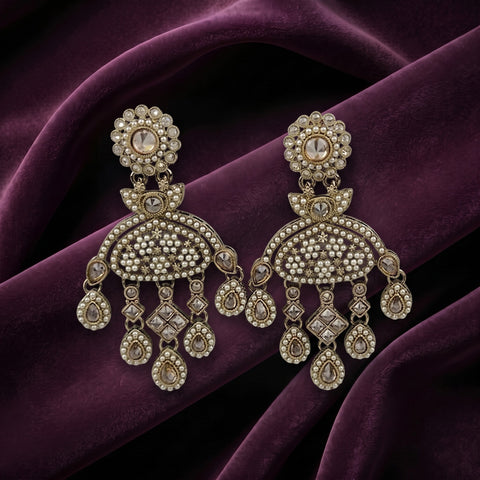 A pair of ornate gold-toned chandelier earrings embellished with clear stones and small pearl-like beads, displayed on draped deep purple fabric.