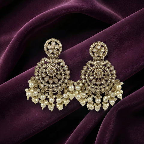 A pair of ornate gold-toned chandelier earrings with intricate filigree work, clear stones, and dangling pearl-like beads, displayed on draped deep purple velvet fabric.