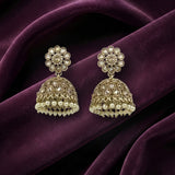 A pair of gold-toned Indian jhumka earrings with clear stone floral studs and dangling pearl-like bead borders, displayed on draped deep purple fabric.