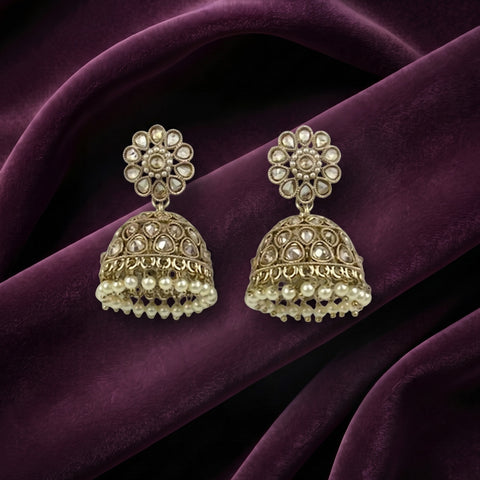 A pair of gold-toned Indian jhumka earrings with clear stone floral studs and dangling pearl-like bead borders, displayed on draped deep purple fabric.