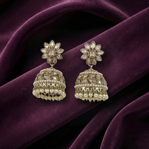 A pair of ornate gold-toned jhumka earrings with floral studs and beaded bell-shaped drops, displayed on draped deep purple velvet fabric.