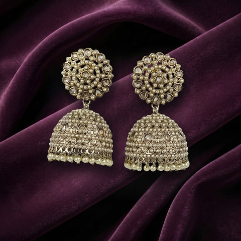 A pair of ornate Indian jhumka earrings with intricate gold-toned filigree and clear stones, displayed on draped deep purple velvet fabric.