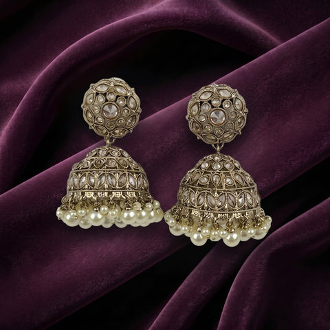 A pair of ornate Indian jhumka earrings with intricate silver-toned filigree work, clear stones, and dangling pearl-like beads, displayed on draped deep purple velvet fabric.