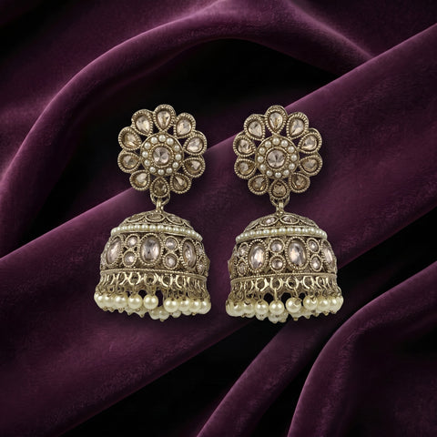 A pair of ornate Indian jhumka earrings with intricate gold-toned filigree, clear stones, and white pearl bead drops, displayed on draped deep purple velvet fabric.
