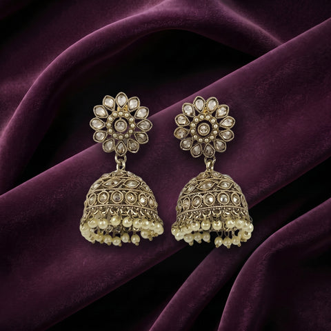 A pair of ornate Indian jhumka earrings with gold-toned filigree, clear stone embellishments, and dangling pearl-like beads, displayed on draped deep purple velvet fabric.