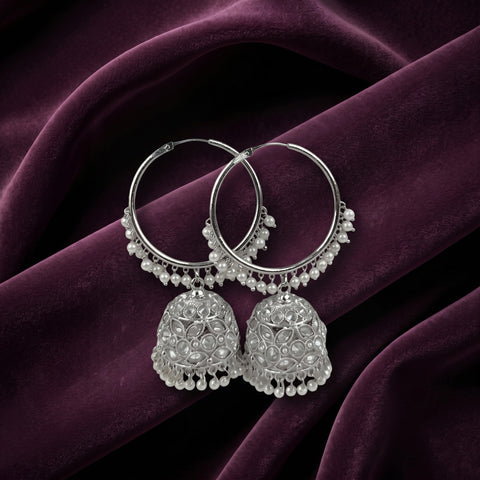 A pair of silver-toned jhumka earrings with intricate beadwork and dangling pearl-like embellishments, displayed on draped deep purple fabric.