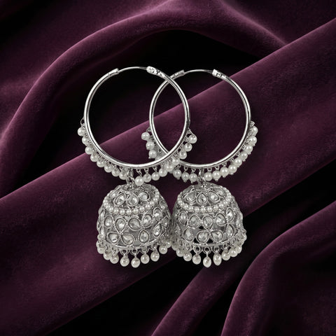 A pair of silver-toned Indian jhumka earrings with intricate beadwork and dangling pearl-like beads, displayed on draped deep purple velvet fabric.