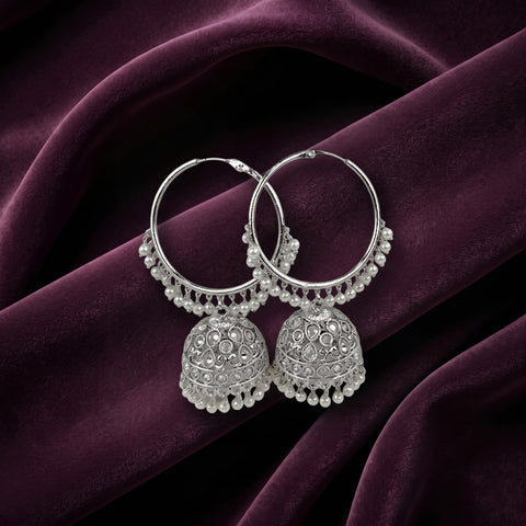 A pair of silver-toned Indian jhumka earrings with intricate filigree work and bead borders, displayed on draped deep purple velvet fabric.