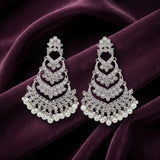 A pair of silver-toned chandelier earrings adorned with clear stones and white pearls, displayed on a draped deep purple velvet fabric.