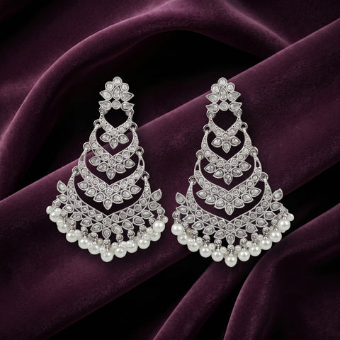 A pair of silver-toned chandelier earrings adorned with clear stones and white pearls, displayed on a draped deep purple velvet fabric.