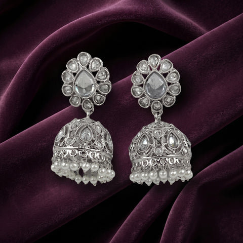 A pair of silver-toned Indian jhumka earrings with clear teardrop stones and pearl bead fringe, displayed on a draped deep purple velvet fabric.