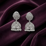 A pair of silver-toned Indian jhumka earrings with intricate filigree work and small clear stones, displayed on draped deep purple velvet fabric.
