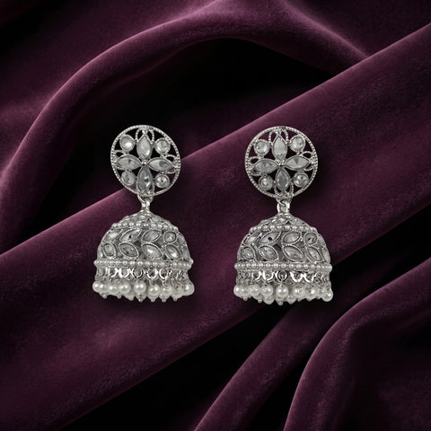 A pair of silver-toned Indian jhumka earrings with intricate filigree work and pearl bead fringe, displayed on draped deep purple velvet fabric.