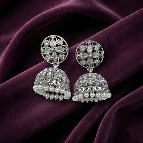 A pair of ornate silver-toned Indian jhumka earrings with floral filigree tops and dangling pearl bead bells, displayed on draped deep purple velvet fabric.