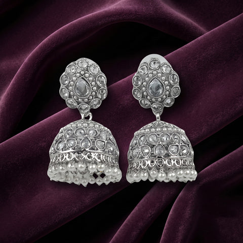 A pair of silver-toned Indian jhumka earrings adorned with clear stones and small pearl-like beads, displayed on a draped dark purple velvet fabric.