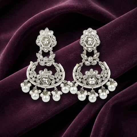 A pair of silver-toned chandelier earrings adorned with clear crystals and white pearls, displayed on a draped deep purple velvet fabric.