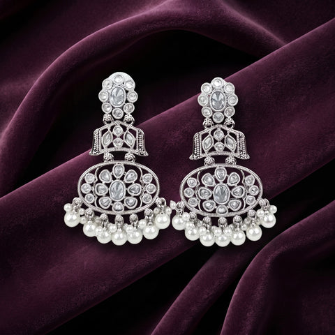 A pair of silver-toned traditional Indian chandelier earrings adorned with clear stones and white pearl drops, displayed on a draped dark purple velvet fabric.