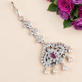 A silver-toned ornate jewelry pendant with clear rhinestones, a central pink gemstone, and dangling white faux pearls, displayed on a light beige background with purple roses in the corner.
