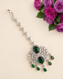 A silver-toned traditional Indian jhumka earring decorated with clear crystals and green emerald-cut gemstones, displayed on a light beige background with a decorative purple rose arrangement in the top right corner.