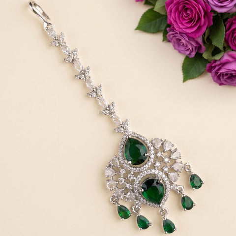 A silver-toned traditional Indian jhumka earring decorated with clear crystals and green emerald-cut gemstones, displayed on a light beige background with a decorative purple rose arrangement in the top right corner.