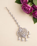A silver-toned Indian style hair ornament (taar) decorated with clear crystals, displayed on a beige background next to a bouquet of purple roses.