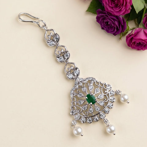 A silver-toned ornate keychain or bag charm with clear rhinestones, a central green stone, and small white pearl drops, photographed on a light beige background with purple roses in the corner.