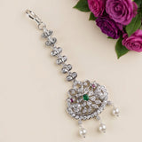 A silver-toned traditional Indian maang tikka with a large floral pendant, embellished with clear crystals, a green center stone, and small red accent stones, with pearl-like dangling beads. The jewelry is displayed on a light beige background next to a cluster of purple roses.