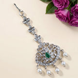 A silver-toned traditional Indian choker necklace with a large ornate pendant, featuring a green teardrop-shaped stone, clear rhinestones, and pearl-like dangling beads, displayed on a beige background with purple roses in the corner.