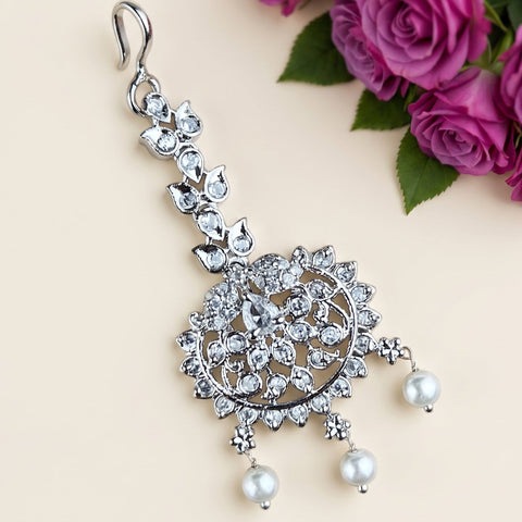 A silver-toned traditional Indian choker necklace adorned with clear crystals and white pearl drops, displayed on a light beige background with purple roses in the corner.