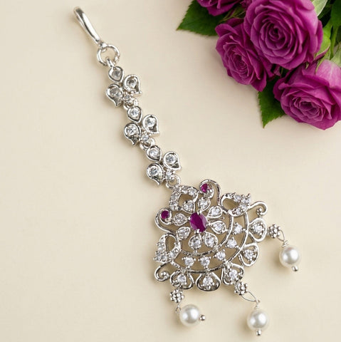 A silver-toned traditional Indian maang tikka with clear crystals, a central purple ruby, and pearl drops, displayed on a beige background with purple roses in the corner.
