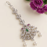 A silver-toned traditional Indian choker necklace with a large ornate pendant, featuring clear stones, a green center stone, two small pink stones, and three dangling white pearls. The necklace is styled on a light beige background with purple roses in the top right corner.