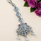 A silver-toned traditional Indian bridal jewelry piece featuring a large floral mandarin design with clear stones, small purple accent stones, and three dangling white pearls, shown next to pink roses on a beige background.