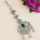 A silver-toned traditional Indian pendant necklace with a green oval-cut stone and white pearl drops, decorated with clear crystals, displayed on a beige background with purple roses in the corner.