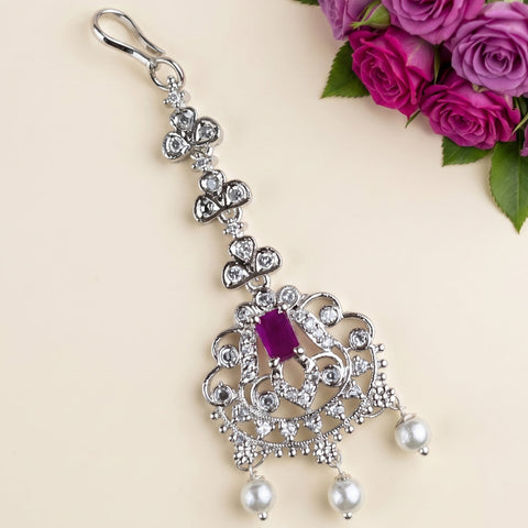 An ornate silver-toned chandelier earring with a central purple gemstone and clear rhinestones, displayed on a light beige background with a bouquet of purple roses in the top right corner.