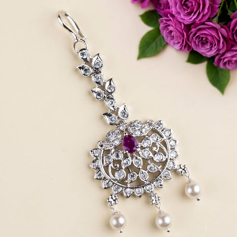 A silver-toned Indian choker necklace with a detailed filigree pendant, featuring a central purple gemstone and three dangling white pearls, styled with pink roses on a beige background.