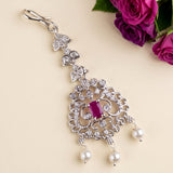 A silver-toned ornate dangle earring decorated with clear crystals, a central rectangular pink gemstone, and three dangling white pearls, shown on a beige background with purple roses in the corner.