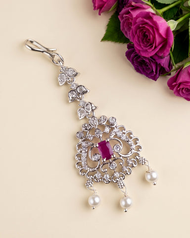 A silver-toned ornate dangle earring decorated with clear crystals, a central rectangular pink gemstone, and three dangling white pearls, shown on a beige background with purple roses in the corner.
