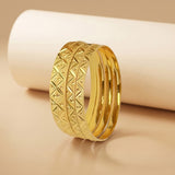 A gold-toned multi-band ring with a textured geometric diamond pattern on the front band, displayed on a beige surface with a white cylindrical prop.