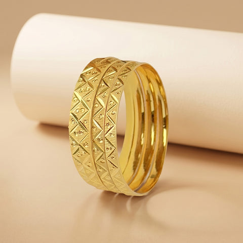 A gold-toned multi-band ring with a textured geometric diamond pattern on the front band, displayed on a beige surface with a white cylindrical prop.