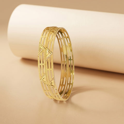 A gold-colored bangle bracelet with an intricate openwork geometric pattern, displayed on a warm beige surface with a matching beige cylindrical prop in the background.